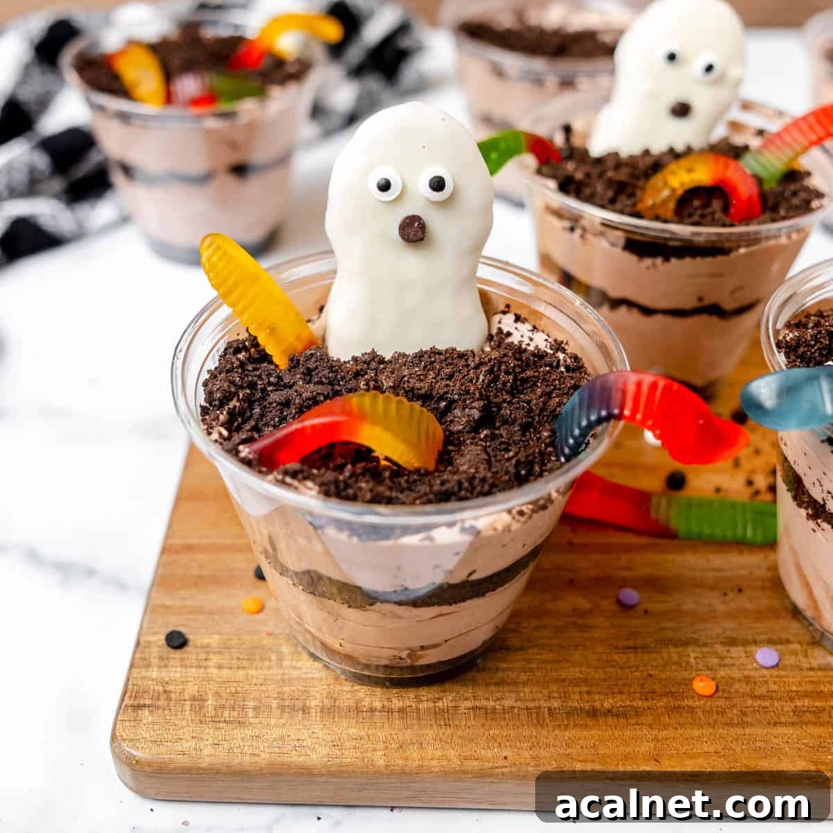 A plastic cup filled with layered chocolate pudding, crushed cookies, gummy worms, and topped with a white chocolate ghost-shaped cookie, displayed on a wooden board.