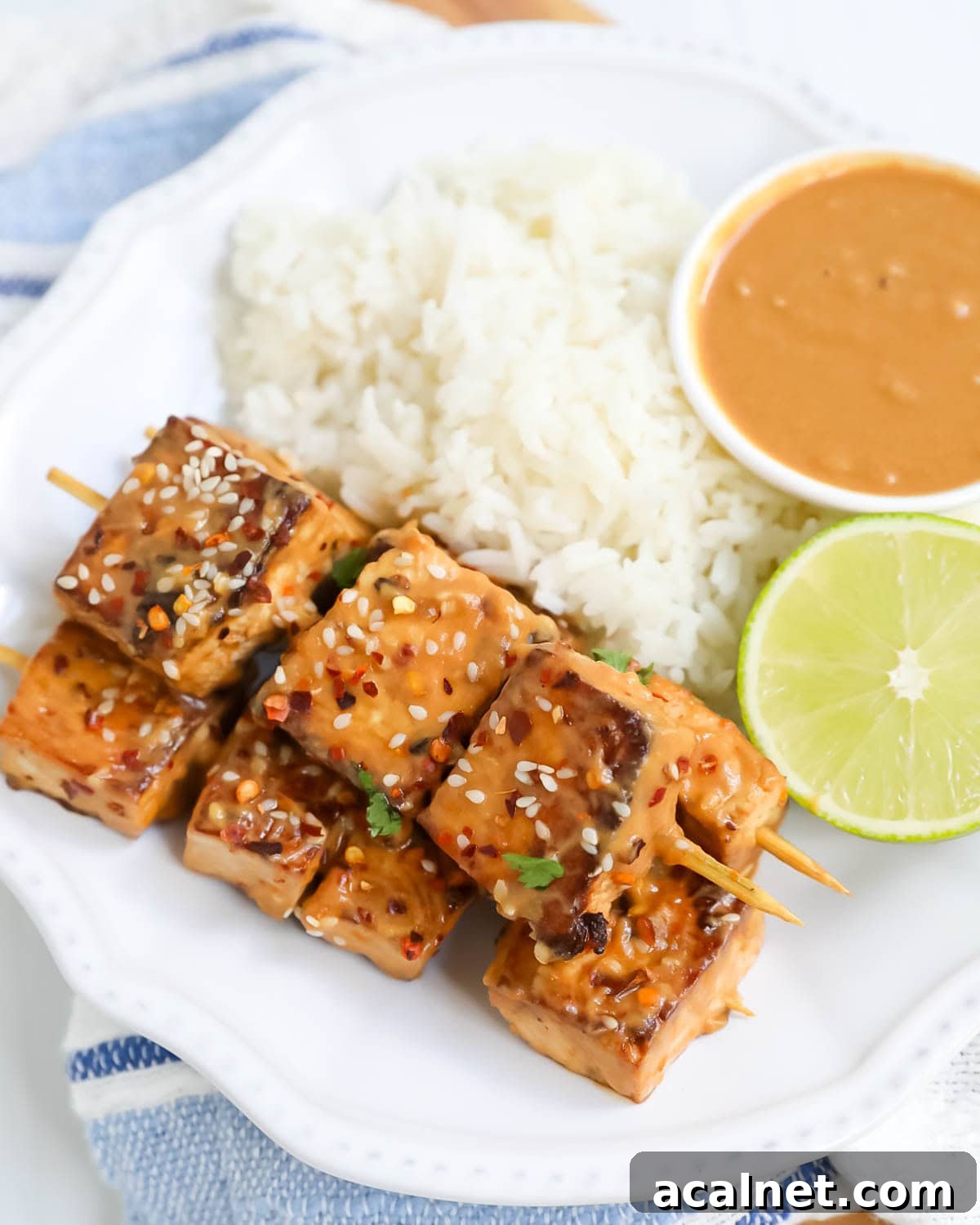 Peanut tofu skewers on a plate with rice, peanut sauce, and fresh limes.