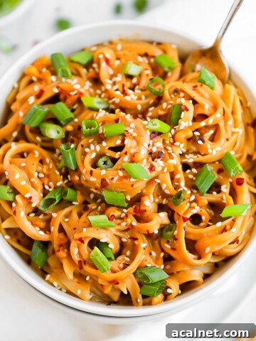 A white bowl filled with creamy peanut butter noodles. They are garnished with green onions and white sesame seeds.