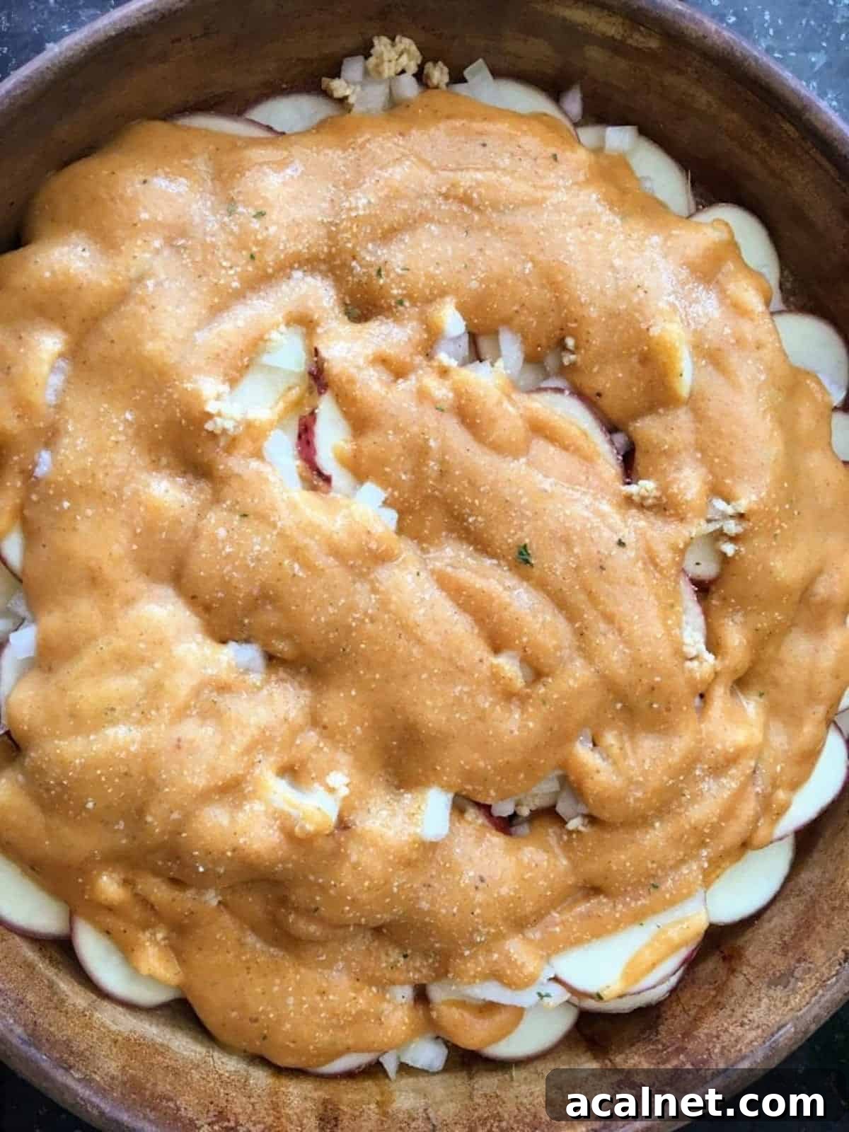 Vegan cheese sauce is poured over the scalloped potatoes recipe, onion, and garlic in a casserole pan. 