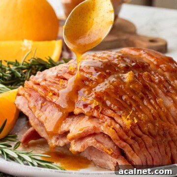Spoon drizzling orange glaze over cooked ham on platter.