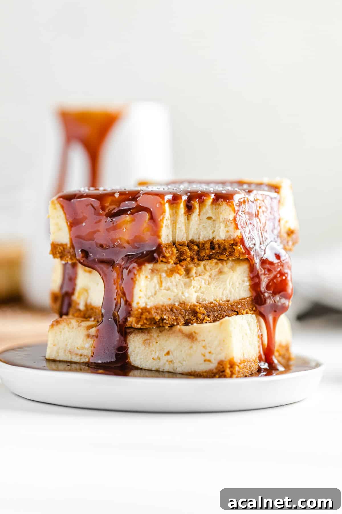 stack of cheesecake bars on a small white plate with a pool of caramel sauce on it and a pitcher of caramel in the background