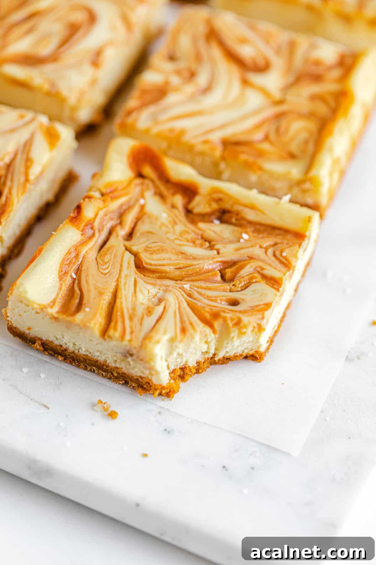 close up of cheesecake bars with caramel swirled on top in rows on a square marble board and a bite taken out the front bar