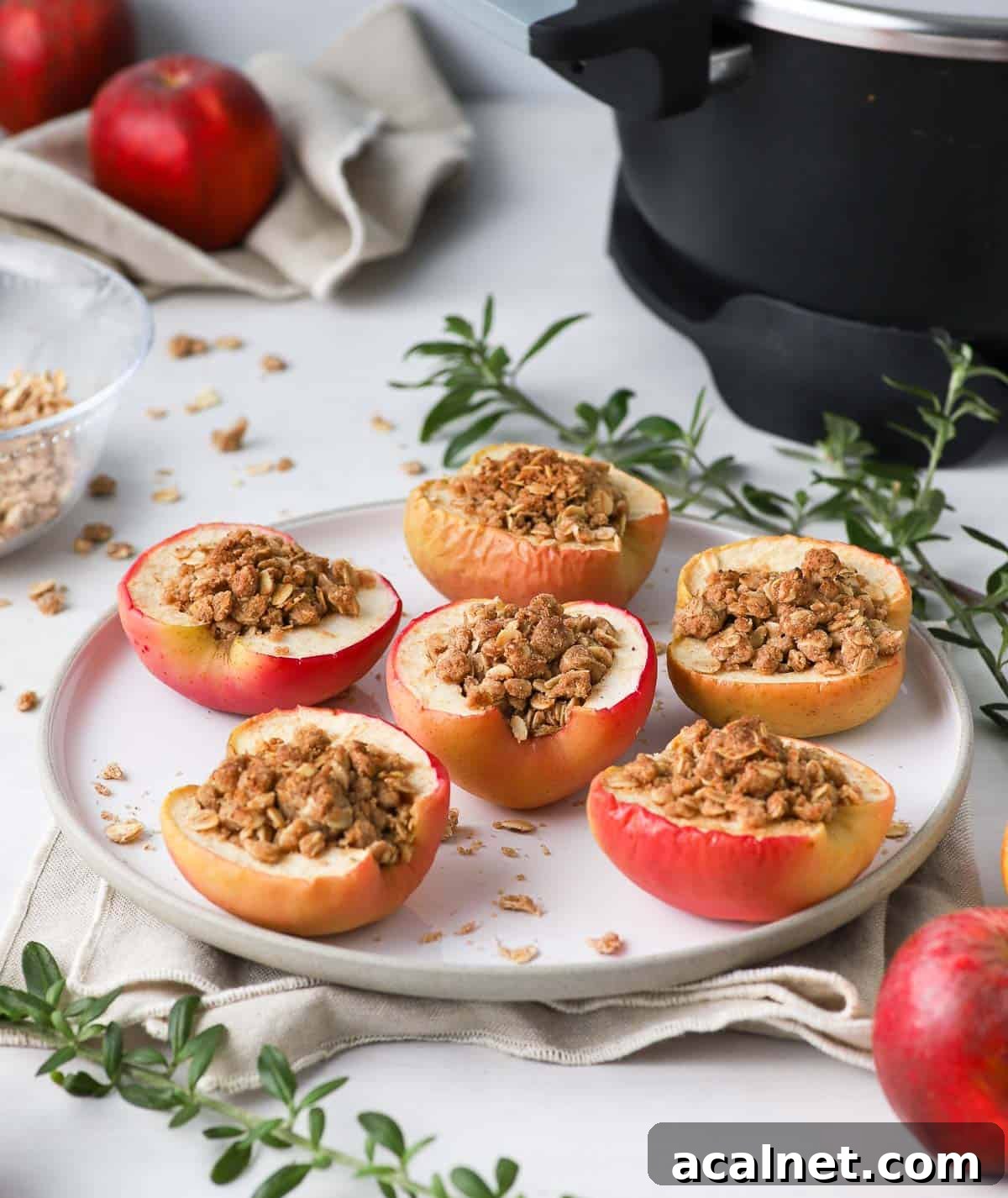 Remoska Baked Apples with Cinnamon Crumble 6 Baked apples on a white plate surrounded by apples, greens and the Remoska.