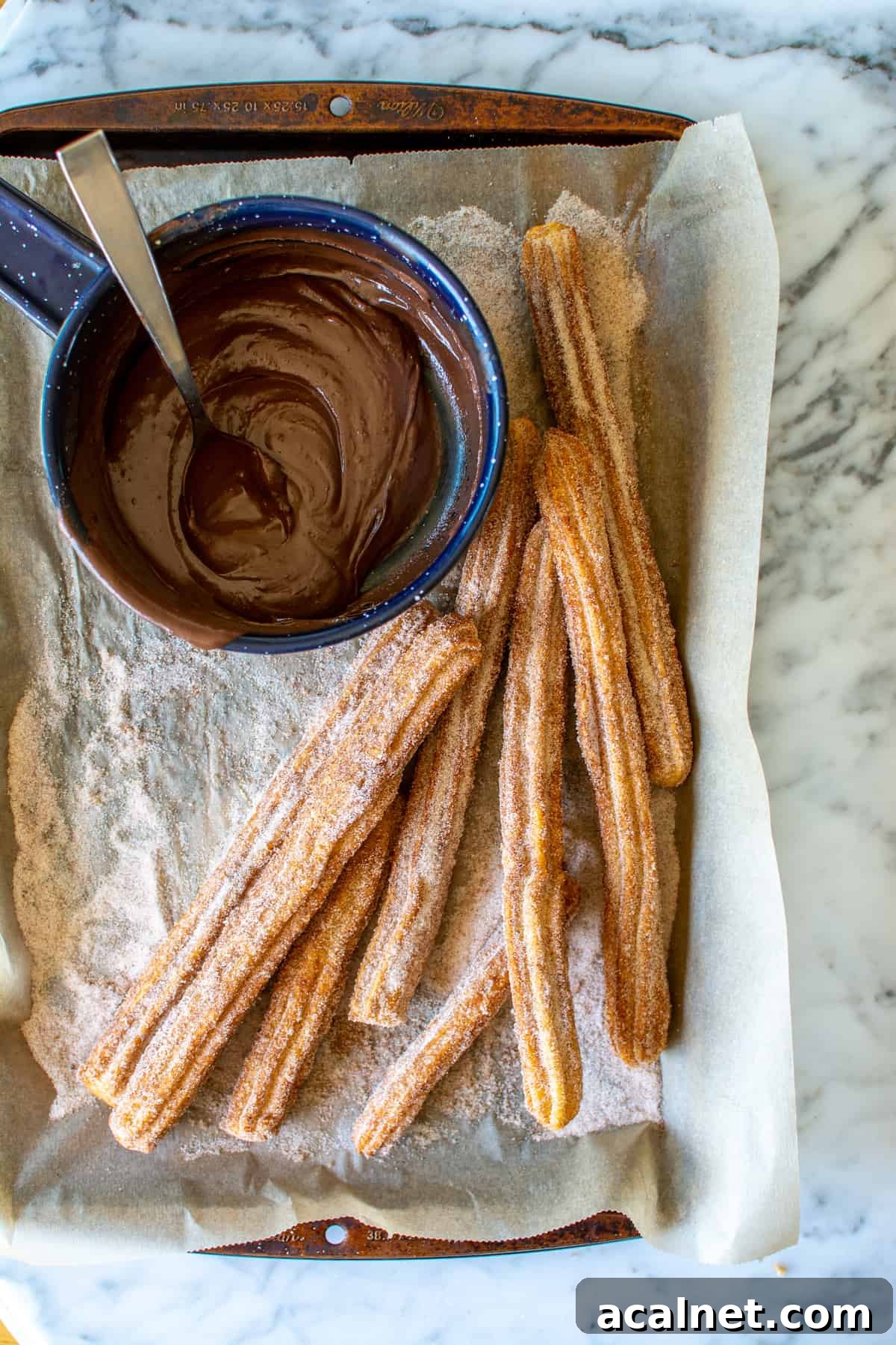 Several churros on a parchment-lined baking sheet with a blue saucepan of chocolate sauce next to them.