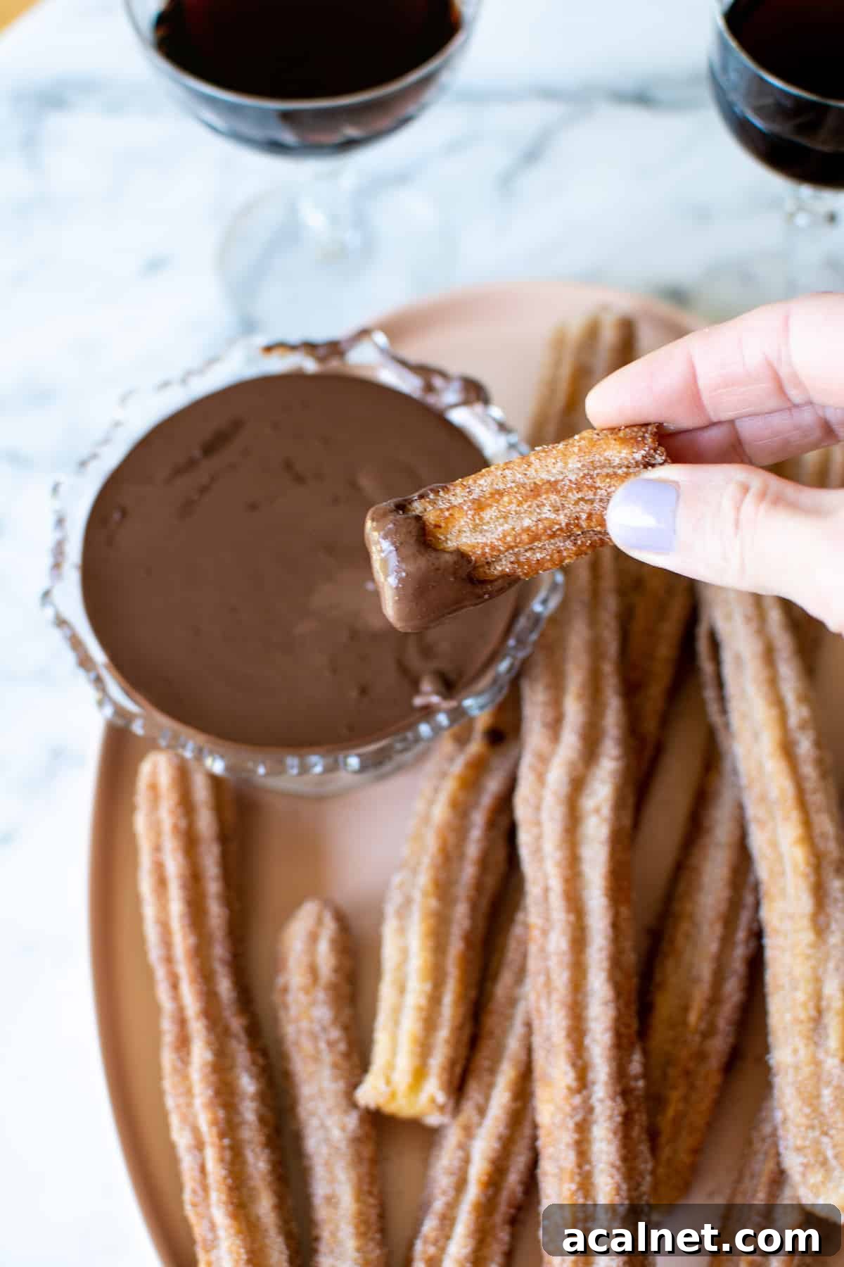 A hand holding a churro that has been dipped in chocolate sauce with a platter of churros and chocolate sauce behind it.