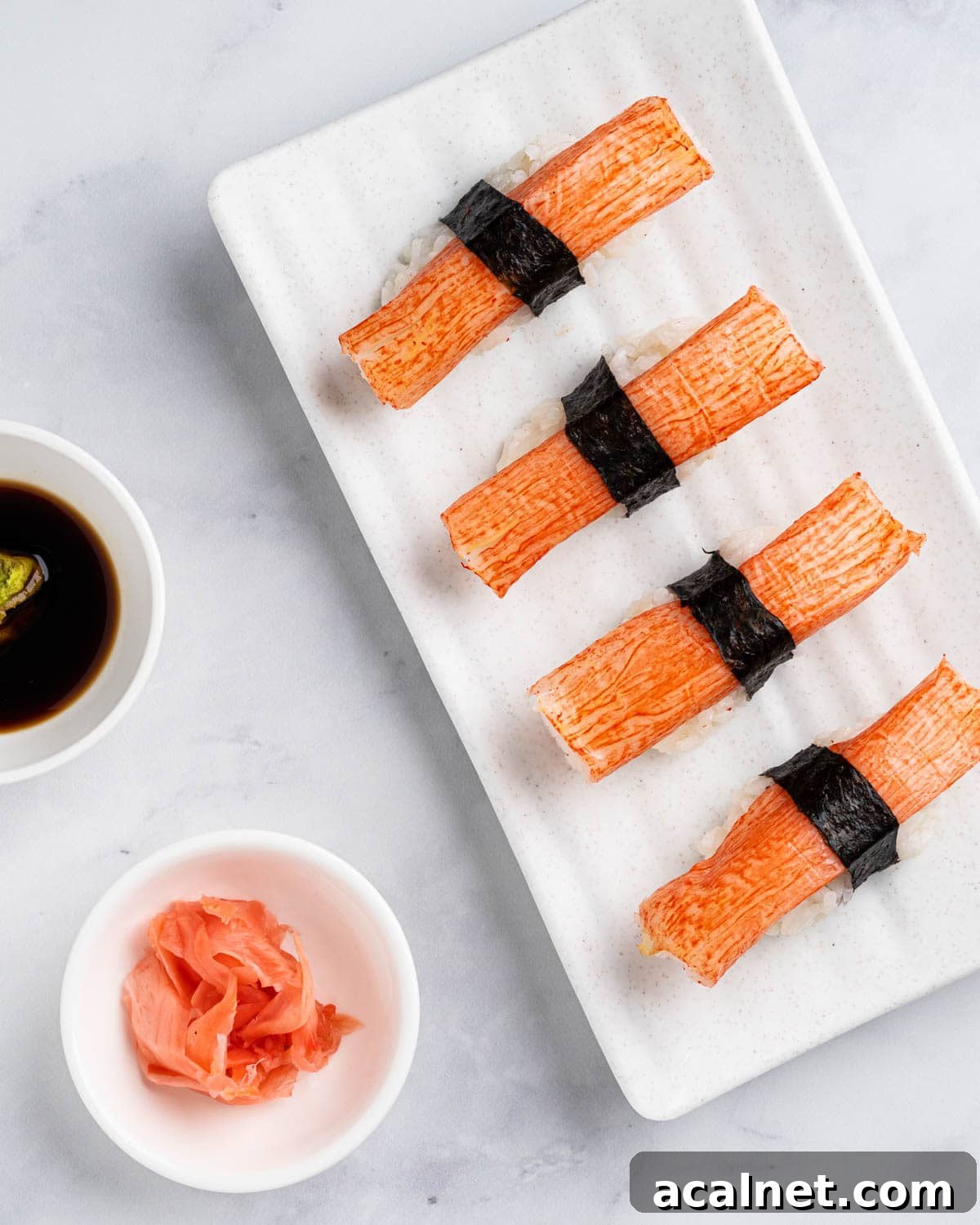 4 Kanikama nigiri sushi on a white plate with a side of soy sauce, wasabi and pickled ginger.
