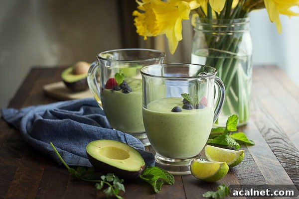 Keto Avocado Smoothie with Cilantro and Mint 3 Green avocado smoothies garnished with berries and mint in glass handled mugs with lime, mint and cilantro placed next to a blue napkin and yellow daffodils.