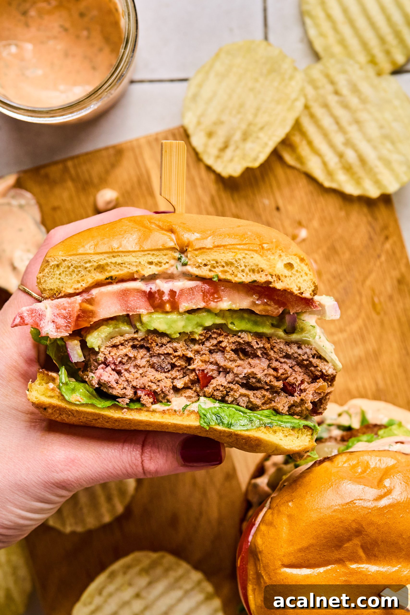 Smoky Chipotle Beef Burgers with Creamy Aioli 12 A person's hand holding a chipotle beef burger with a bite taken out of it surrounded by a jar of homemade chipotle aioli and crinkle potato chips