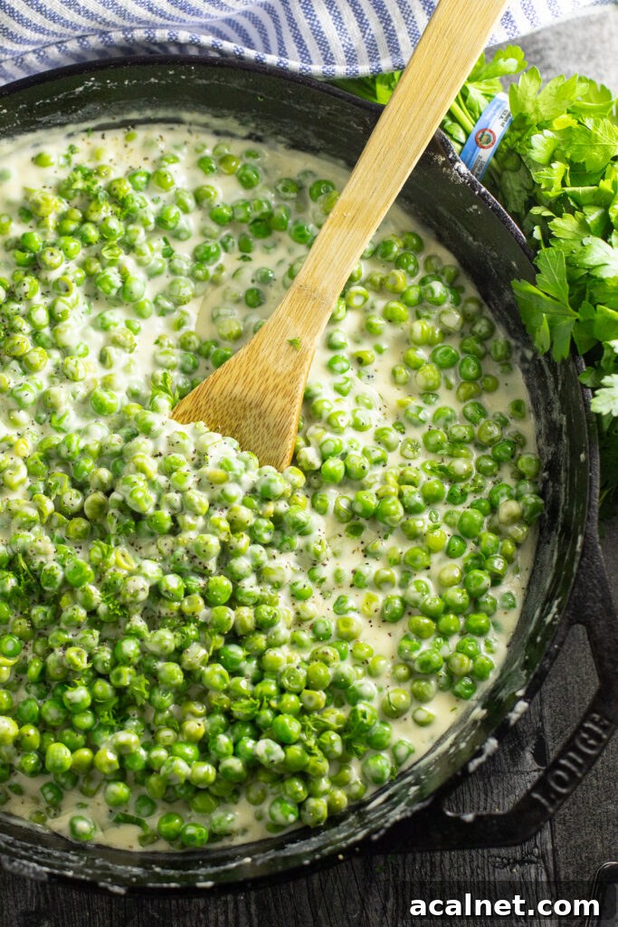 Overhead view of a cast iron skillet filled with creamed peas with a wooden spoon in the center, surrounded by a bunch of fresh parsley and a striped linen napkin.