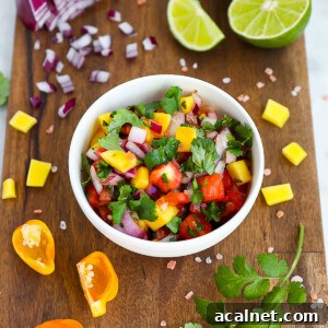 A white bowl sitting on a dark wooden cutting board, the bowl is filled with bright and colorful homemade mango salsa. Ingredients from the salsa are scattered on the cutting board.