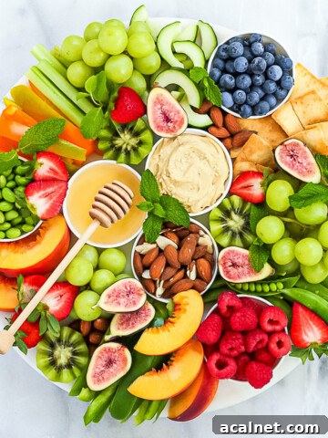 A white marble board filled with fresh fruits, vegetables, hummus, nuts, agave nectar, and garnished with fresh mint leaves.