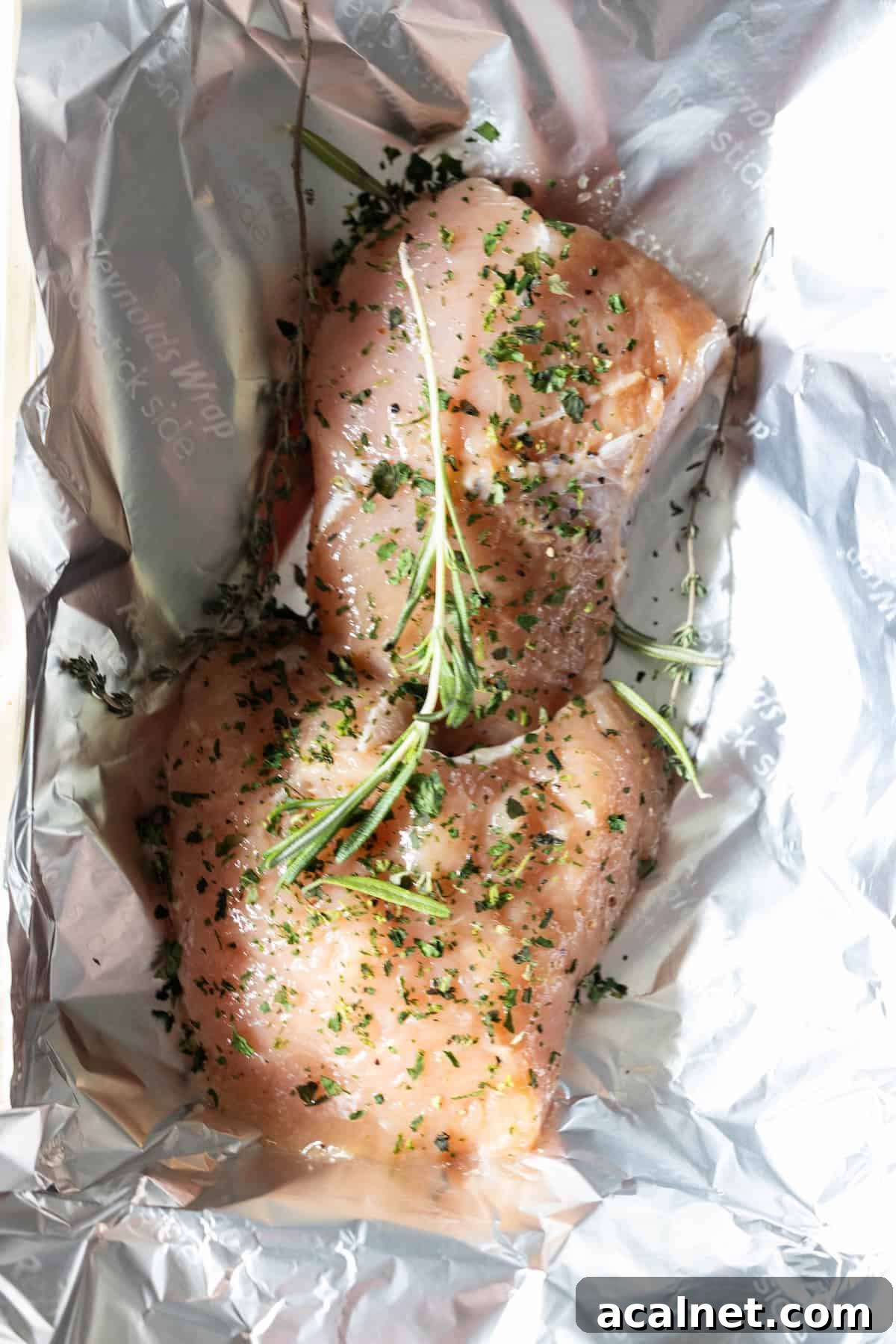 Raw chicken breasts seasoned with herbs and a sprig of rosemary, placed on a sheet of aluminum foil—just like preparing an easy turkey recipe or baking turkey tenderloin for a flavorful meal.