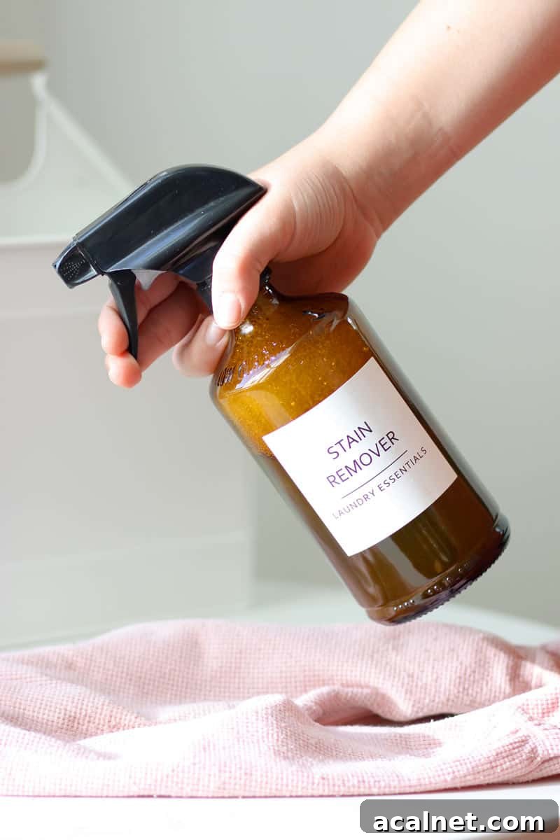 Homemade Laundry Stain Remover Spray Recipe 2 a glass amber spray bottle with a label that says 'stain remover laundry essentials' on it being sprayed at a stained baby outfit