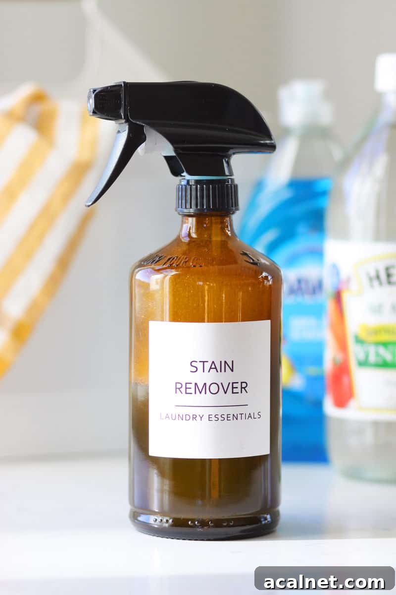 Homemade Laundry Stain Remover Spray Recipe 5 a glass amber spray bottle with a label that says 'stain remover laundry essentials' and a bottle of dawn dish soap and a bottle of distilled vinegar behind it