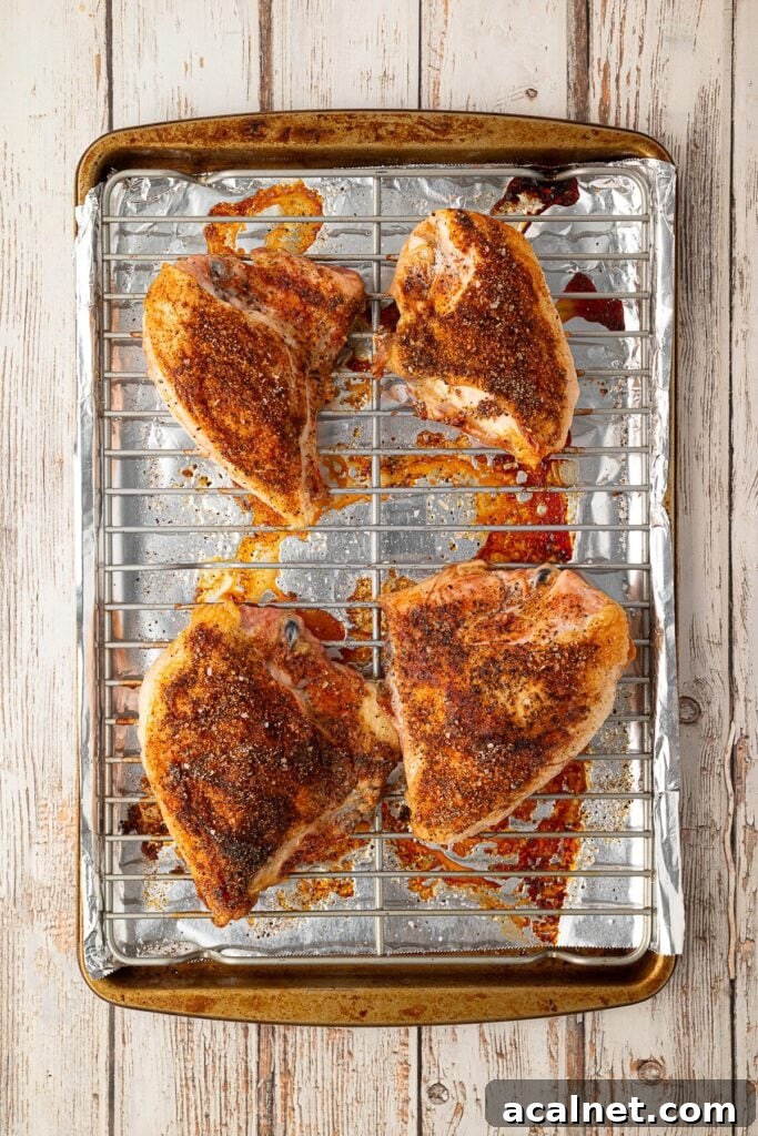 Freshly baked seasoned split chicken breasts resting on a baking sheet with crispy skin and clear pan juices.