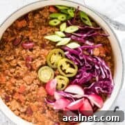 Venison chili in a white bowl, topped with green onion, tomatoes, cabbage, and jalapeno slices.