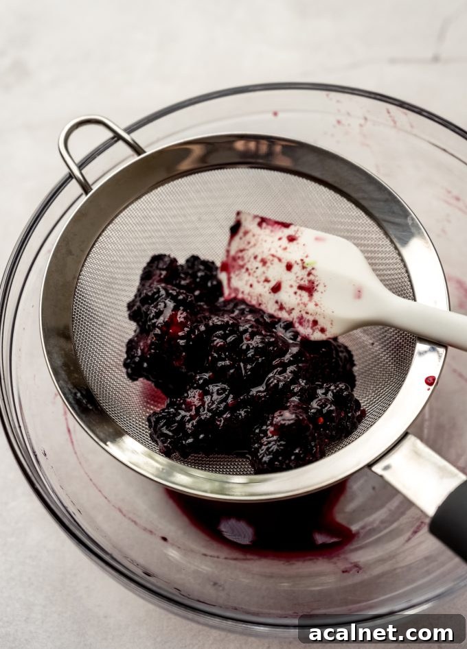Someone is using a spatula to strain macerated blackberries through a mesh sieve.