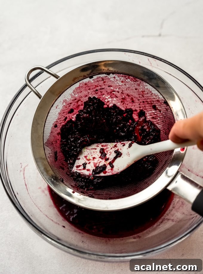 Someone is using a spatula to strain macerated blackberries through a mesh sieve.