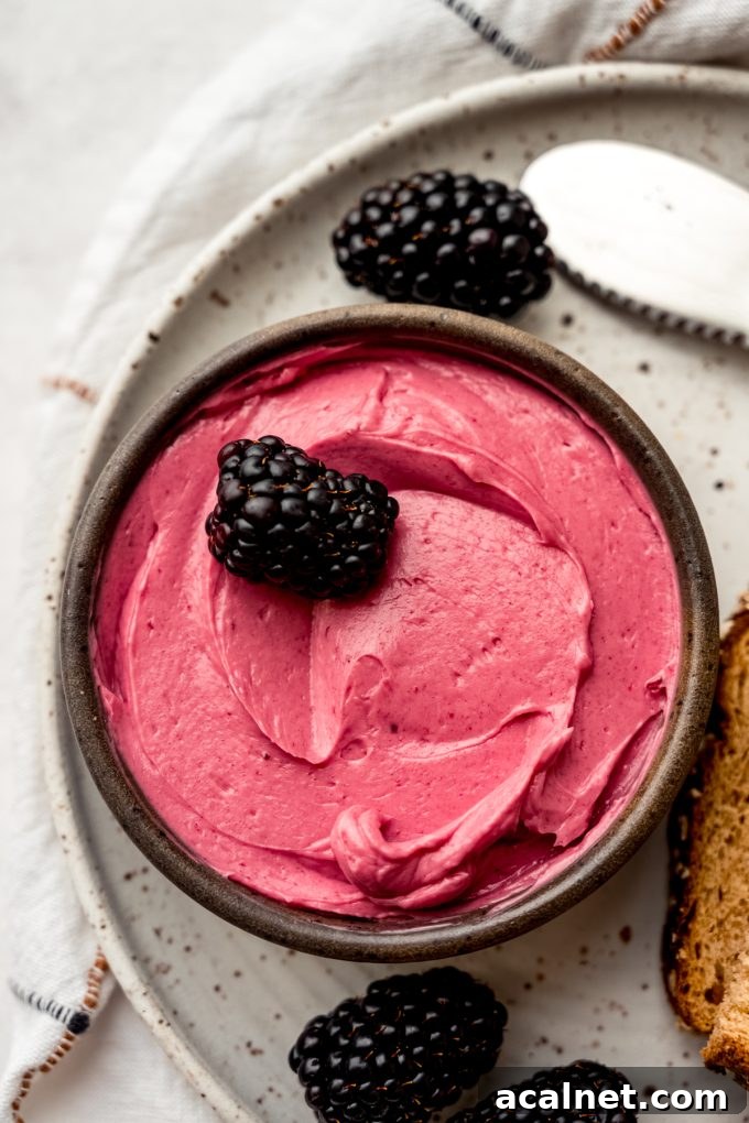 A small bowl of blackberry butter on a plate with a blackberry in it and other blackberries around the plate.